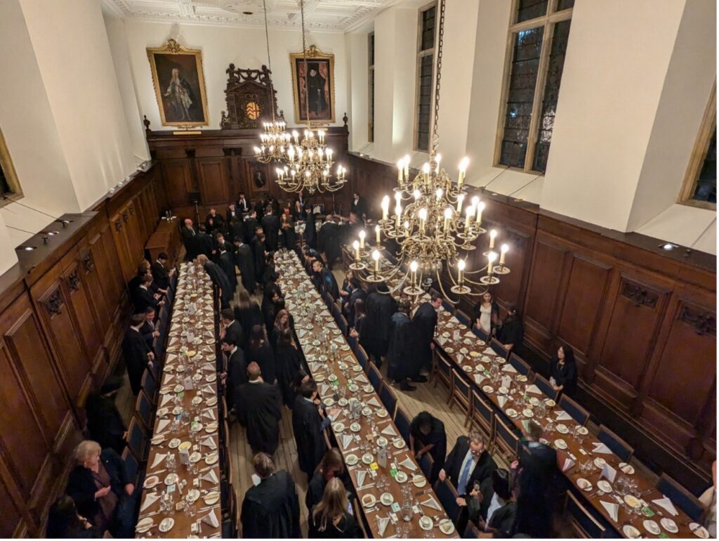 ケンブリッジ大学のフォーマルホールで、ローブ姿の学生たちが長テーブルに着席している様子。